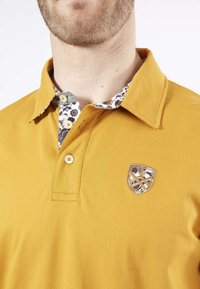 Polo shirt jaune avec un col à motifs, présentant des designs floraux bleus. Il dispose d'un seul bouton et d'un logo en broderie en forme de blason sur la poitrine.