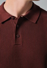 Polo en côte bordeaux avec un col rabattu, trois boutons et un tissu texturé. Gros plan mettant en avant le motif de tricot.