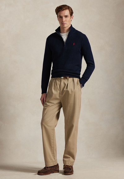 Młody mężczyzna ubrany w granatowy sweter z zamkiem do połowy długości, beżowe spodnie z plisami i brązowe skórzane buty, stojący na tle jednolitego beżowego tła.