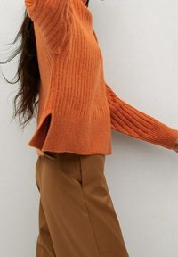 Pull orange à côtes avec des manches longues et des fentes sur les côtés, associé à un pantalon marron. Le pull présente un design tricoté texturé.