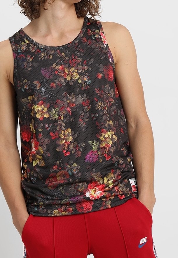 Jeune personne portant un haut noir sans manches à motif floral et un pantalon rouge Nike, les mains dans les poches, sur un fond uni.