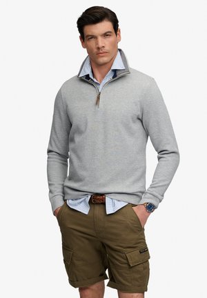Mand iført lysegrå quarter-zip sweater over blåstribet skjorte, brune cargoshorts, flettet bælte og armbåndsur, stående med hænderne i lommerne.
