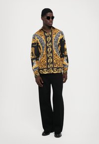 Bărbat îmbrăcat cu ochelari de soare și pantaloni negri, purtând o cămașă cu un model negru și auriu, decorată cu tigri și pete de leopard.