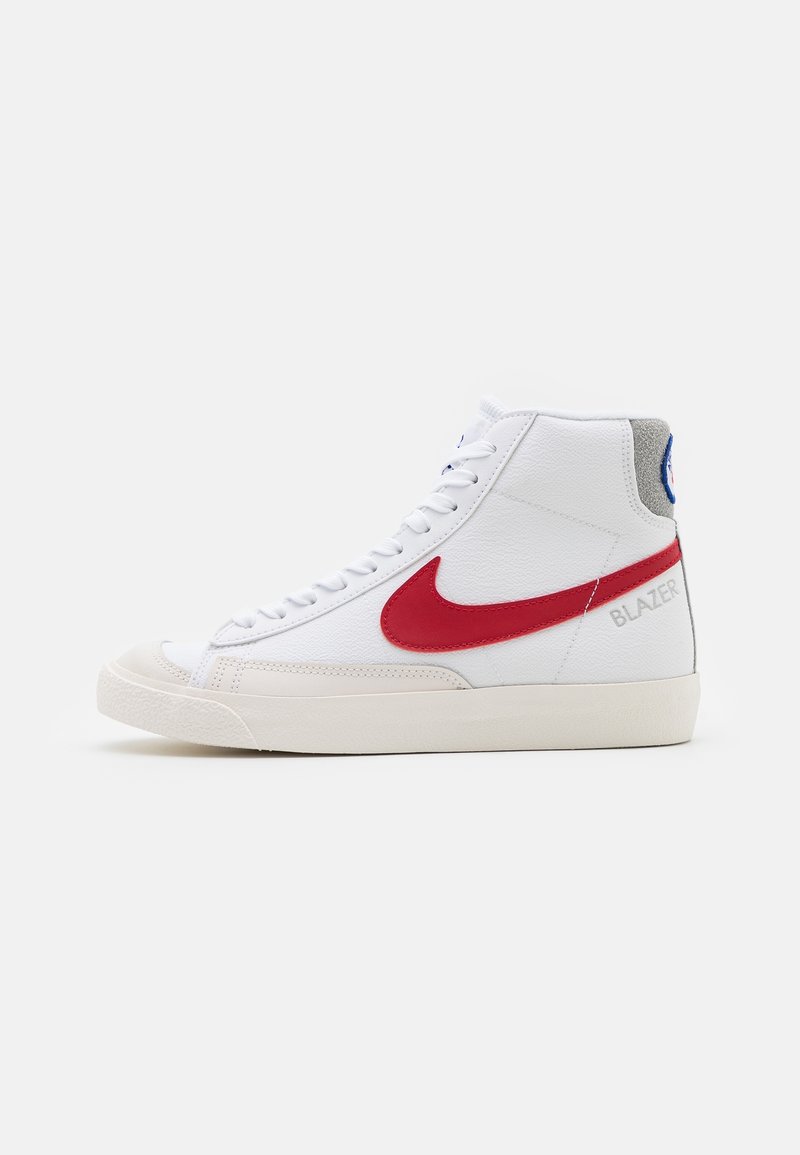 Zapatilla alta blanca con el logo de Nike en rojo, suela beige, punta redonda y el texto "BLAZER" cerca del talón sobre un fondo blanco liso.