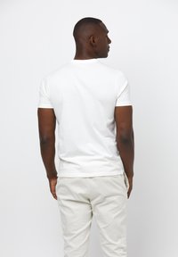 T-shirt en coton blanc à manches courtes, de forme ajustée et avec un col rond. Le modèle porte un pantalon de couleur claire, vu de dos.