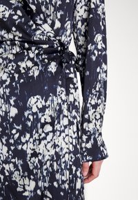 Zavinovacia šaty z námorníckej látky s bielym abstraktným vzorom. Detailné dlhé rukávy s gombíkovými manžetami. Viazaný pás pre prispôsobiteľný strih.