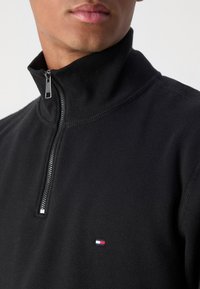 Gros plan d'une personne portant un pull noir zippé avec un petit logo rouge, blanc et bleu sur la poitrine.