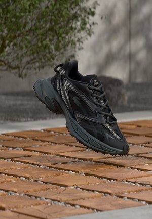 Baskets de sport noires avec des accents gris, suspendues au-dessus de carreaux en briques orange à l'extérieur, près d'un mur en béton et de verdure.