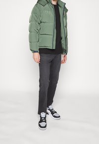 Veste matelassée verte avec capuche, fermeture éclair, et accents bleu marine, associée à un jean foncé et des baskets noires et blanches.