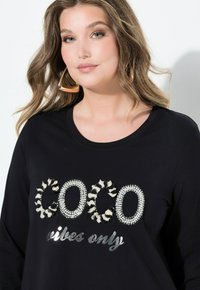 Schwarzes Langarmshirt mit dem Schriftzug "COCO" aus glänzenden Strasssteinen und einem Leopardenmuster-Akzent, dazu der Text "vibes only" in silberner Metallicschrift.