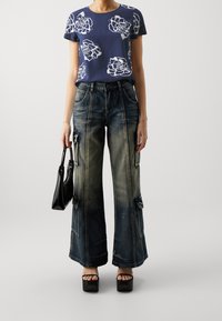 Kvinna i en marinblå blommig t-shirt, blekta vida cargobyxor, svarta plattformssandaler, och håller en handväska i svart lackläder.