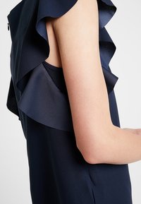 Close-up van de arm van een persoon die een mouwloos marineblauw topje draagt met gelaagde ruches bij de schouder.