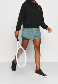 Schwarzer Kapuzenpullover, strukturierte türkisfarbene Shorts und schwarze Sportschuhe. Hält einen weißen Tennis-Schläger und zeigt ein lässiges sportliches Outfit.