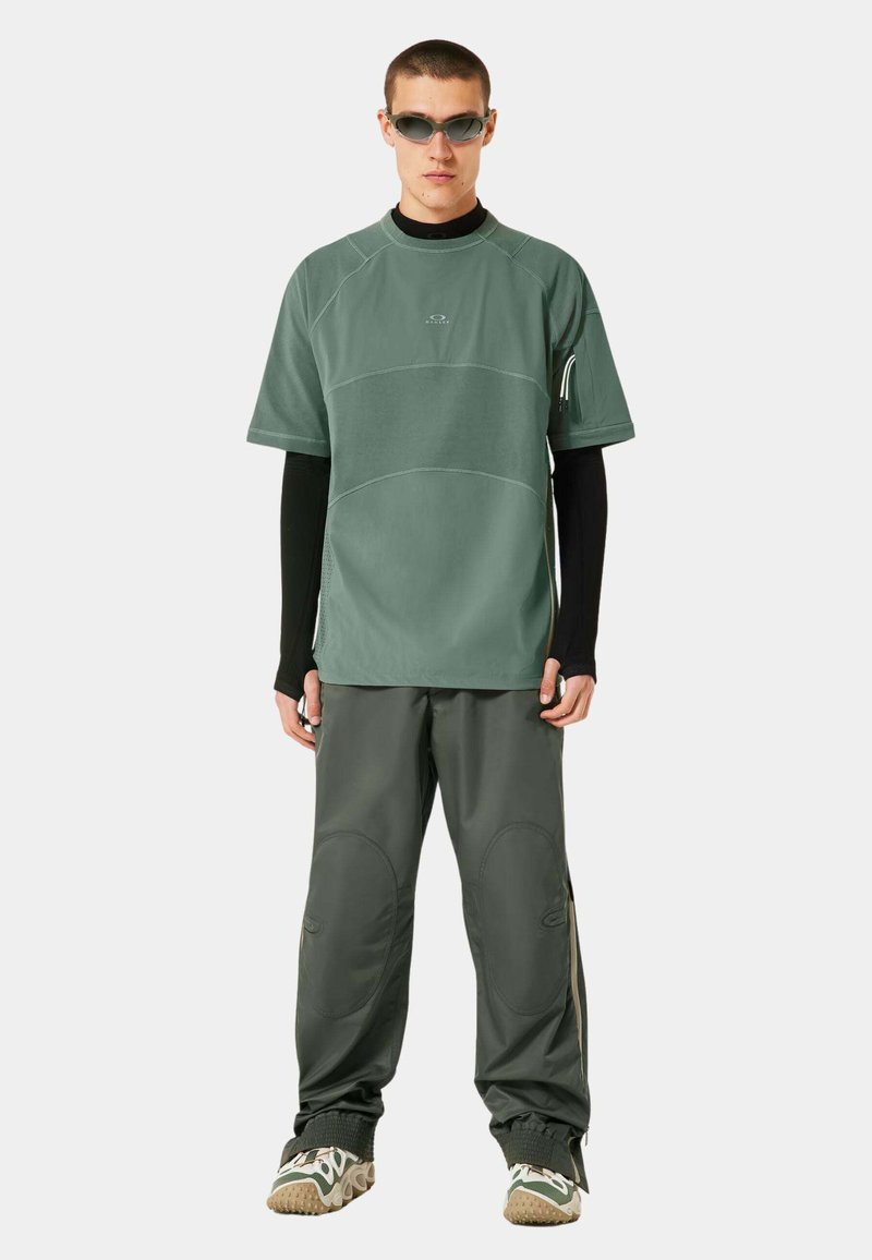 Haut vert à manches courtes avec des détails de couture, associé à un pantalon vert foncé avec des poches latérales et des accents texturés. Porté avec des lunettes de soleil.