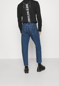 Černá košile s dlouhým rukávem a bílým nápisem "Calvin Klein Jeans" na zadní straně, v kombinaci s tmavě modrými zúženými džíny a černými botami.