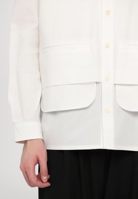 Hvit skjorte med to frontlommer, knapp-up design og lange ermer. Materialet virker glatt med en lett tekstur.