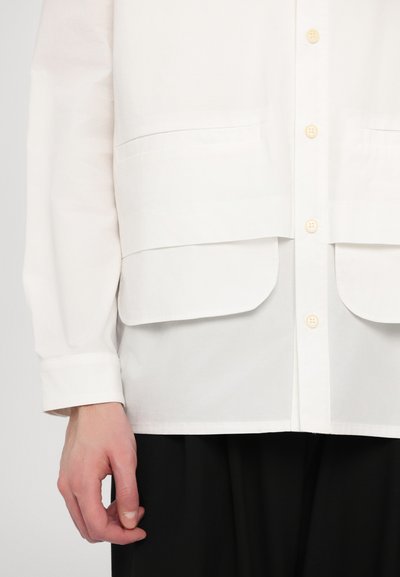 Camisa blanca con dos bolsillos frontales, diseño abotonado y mangas largas. El material parece suave con una textura ligera.