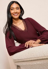 Bordeauxfarbenes Oberteil mit einem Spitzen-V-Ausschnitt, strukturiertem Stoff und langen, puffigen Ärmeln. Das Model lehnt die Hände auf einen weichen, hellfarbenen gepolsterten Stuhl.