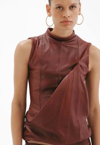 Top marrón de cuero sin mangas con cuello alto, con detalle drapeado en la parte frontal y costuras sutiles, textura suave. Pendientes de aro dorados.