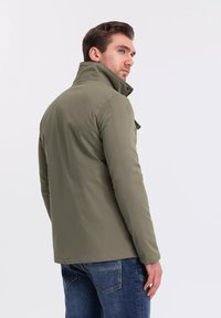 Giacca leggera verde oliva con colletto alto, texture liscia e dettagli con bottoni, abbinata a jeans blu in denim.