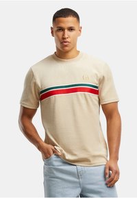 Camiseta beige de manga corta con franjas horizontales en rojo, verde y blanco en el pecho; presenta un cuello redondo y un logo en la parte superior izquierda.