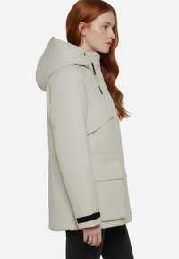 Giacca impermeabile beige chiaro con cappuccio, dotata di tasche frontali, vestibilità leggermente oversize e dettagli in metallo neri.