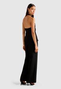 Woman wearing a black halter-back top and wide-leg black trousers, standing and looking over her shoulder toward the camera.