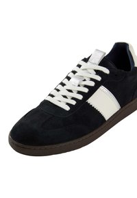 Sneaker in suede nera con lacci bianchi, accenti argentati e una striscia laterale bianca; presenta un colletto imbottito e una suola in gomma con texture.