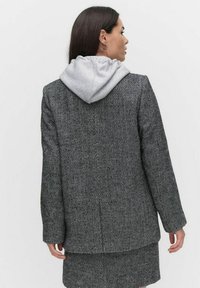 Mustapohjainen herringbone-kuvioitu takki harmaalla hupulla; sisältää rakenteelliset olkapäät, pitkät hihat ja muokatun istuvuuden.