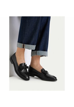 Füße in schwarzen Leder-Loafers mit Schleifendetail, kombiniert mit dunkelblauen, aufgekrempelten Denim-Jeans vor einfachem hellem Hintergrund.