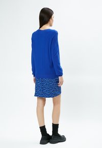 Langärmliger blauer Pullover mit strukturierten Schultern, kombiniert mit einem kurzen, gemusterten Rock in Blau und Weiß, getragen mit schwarzen Socken und Plateauschuhen.