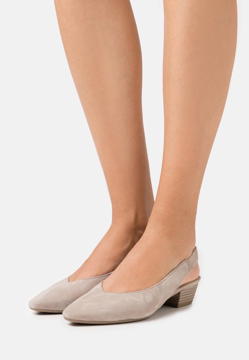 Jambes portant des chaussures slingback en suédine gris clair avec des talons empilés bas sur fond blanc.