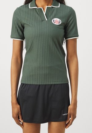 Vrouw draagt een groene geribde golfpolo met een klein logo en een zwarte sportrok met een merknaam op de zoom.