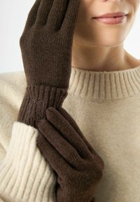Braune Strickhandschuhe mit gerippten Bündchen und einem Schmetterlingsstickdetail, getragen zu einem cremefarbenen gerippten Pullover. Nahaufnahme.