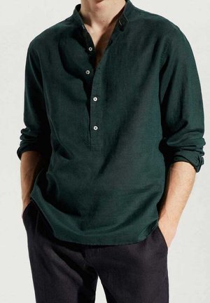 Mann trägt ein dunkelgrünes Henley-Shirt mit langen Ärmeln und drei Knöpfen, Ärmel hochgekrempelt, Hände in den Taschen, vor einem einfarbigen Hintergrund.
