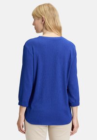 Haut en tricot bleu texturé avec des manches trois-quarts, encolure ronde et un ourlet légèrement courbé. Coupe décontractée avec un subtil détail en côtes.