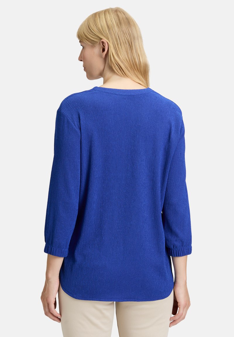 Haut en tricot bleu texturé avec des manches trois-quarts, encolure ronde et un ourlet légèrement courbé. Coupe décontractée avec un subtil détail en côtes.