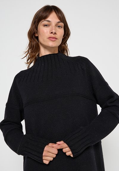 Femme à la peau claire et aux cheveux bruns portant un pull noir côtelé à col haut, les mains jointes à la taille.