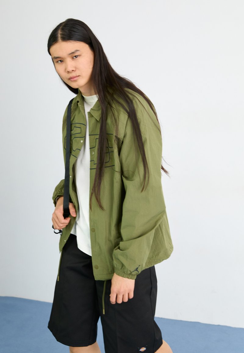 Giacca leggera verde oliva con chiusura a bottoni e design del logo, abbinata a una maglietta bianca e pantaloni corti neri. Tessuto liscio, vestibilità rilassata.
