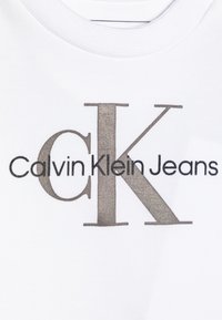 Bijela pamučna majica s istaknutim sivim logom "Calvin Klein Jeans" s stiliziranim "CK" u teksturiranom fontu.