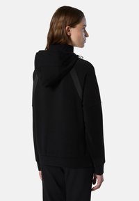 Haut à capuche noir avec cordon de serrage, coupe décontractée, poignets côtelés et deux panneaux latéraux texturés. Tissu doux avec une finition lisse.