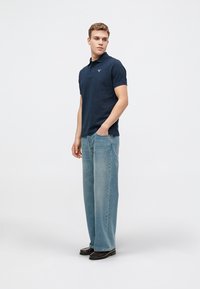 Ναυτικό κοντομάνικο πουκάμισο πόλο με διακριτό λογότυπο, συνδυασμένο με ανοιχτό μπλε παντελόνι σε φαρδιά γραμμή και μαύρα παπούτσια, σε ουδέτερο φόντο.