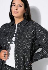 Schwarzes, glitzerndes Oversized-Shirt mit zwei Brusttaschen, Knopfleiste und strukturiertem Finish. Getragen über einem weißen T-Shirt mit Grafik.