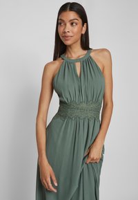 O rochie fără mâneci, de culoare verde măsliniu, cu decolteu în formă de lacăt și talie accentuată cu dantelă, realizată dintr-un material moale și fluid.