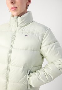 Ljusgrön puffjacka med hög krage, dragkedjestängning, sidfickor och en liten logotyp på bröstet. Slät, glänsande yta.