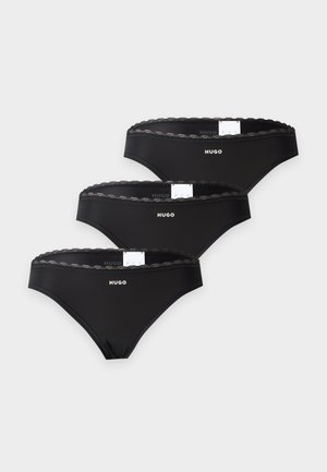 Femme aux longs cheveux foncés portant un soutien-gorge noir et des sous-vêtements noirs avec le logo "Hugo" sur fond blanc.