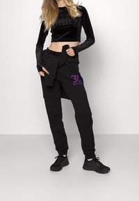 Černý sametový crop top s dlouhým rukávem a reliéfním logem "Juicy", spárovaný s černými tepláky s fialovým logem a elastickým pasem. Černé tenisky.