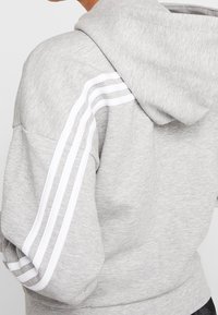 Šedá mikina s měkkou texturou, se bílými pruhy podél rukávů a stahovací kapucí. Jednoduchý, sportovní design.