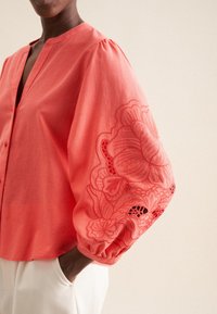 Koralowa bluzka z dekoltem w kształcie litery V i guzikami, z wyszywanymi kwiatowymi wzorami na obszernej bawełnianej części rękawów i teksturowanym materiałem.