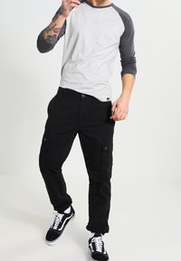 Persoon gekleed in zwarte cargobroeken, zwart-witte sneakers en een grijs shirt met raglanmouwen, staand tegen een vlakke achtergrond.